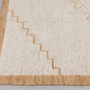 May include: A white rug with a brown border and a geometric pattern in the center. The rug is made of natural fibers and has a textured surface.