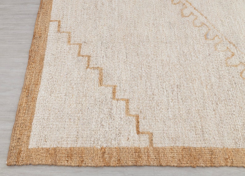 May include: A white rug with a brown border and a geometric pattern in the center. The rug is made of natural fibers and has a textured surface.