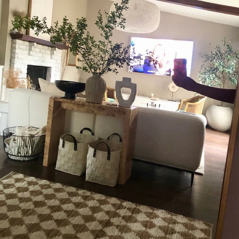 May include: A white couch with a wooden console table behind it. The console table has three woven storage baskets on the bottom shelf. The console table is decorated with a white ceramic vase, a brown ceramic vase, and a white ceramic sculpture. The couch is in front of a large flat screen TV.