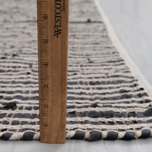 Puede incluir: Primer plano de una alfombra tejida en blanco y negro con una regla de madera que mide 15 cm. La alfombra est&aacute; hecha de tiras de cuero y tela.