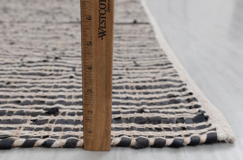 Puede incluir: Primer plano de una alfombra tejida en blanco y negro con una regla de madera que mide 15 cm. La alfombra est&aacute; hecha de tiras de cuero y tela.