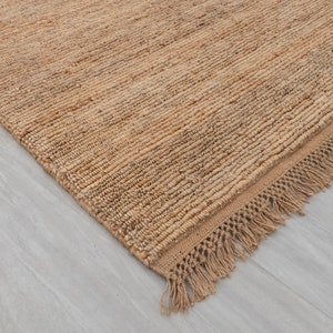 Puede incluir: Primer plano de una alfombra de fibra natural con borde de flecos. La alfombra es de color marrón claro y tiene una textura tejida.