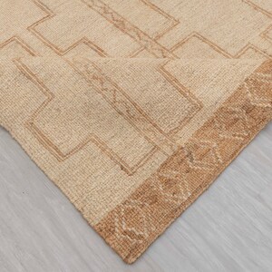 May include: A beige rug with a geometric pattern in a darker beige color. The rug has a brown fringe border.