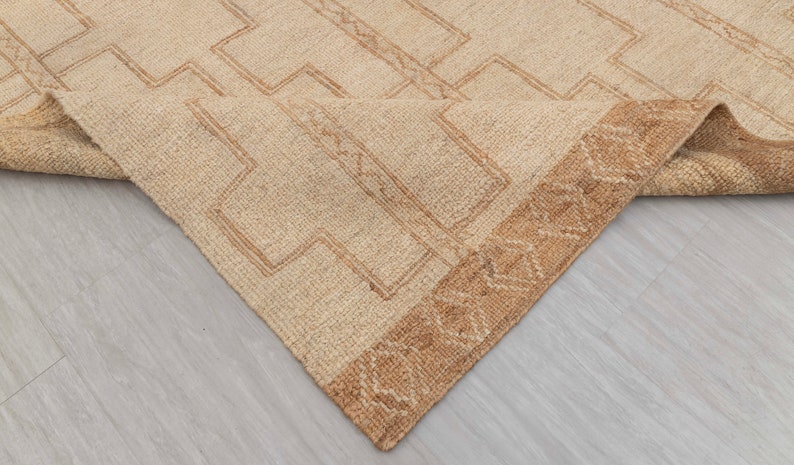 May include: A beige rug with a geometric pattern in a darker beige color. The rug has a brown fringe border.