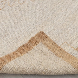 May include: Close-up of a cream-colored rug with a brown geometric pattern. The rug is folded in half, showing the texture of the fabric.