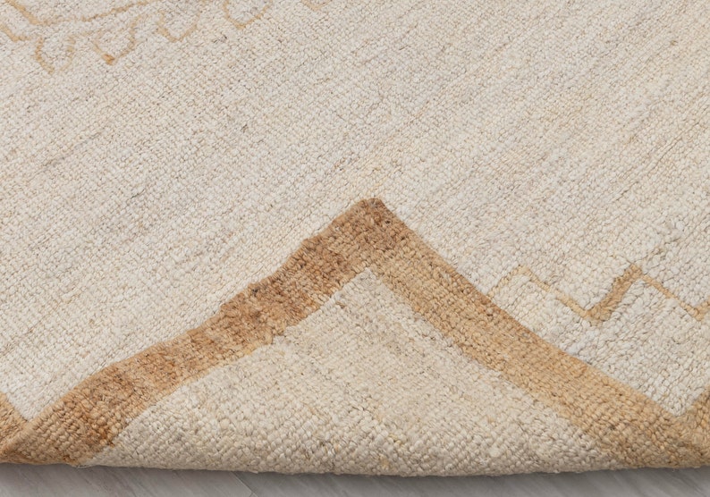May include: Close-up of a cream-colored rug with a brown geometric pattern. The rug is folded in half, showing the texture of the fabric.
