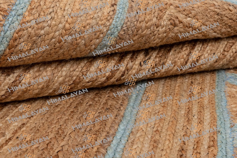 May include: Close-up of a natural fiber rug with a woven pattern. The rug is made of jute and has a light blue stripe running through it. The rug is folded in thirds, showing the texture and weave of the material.