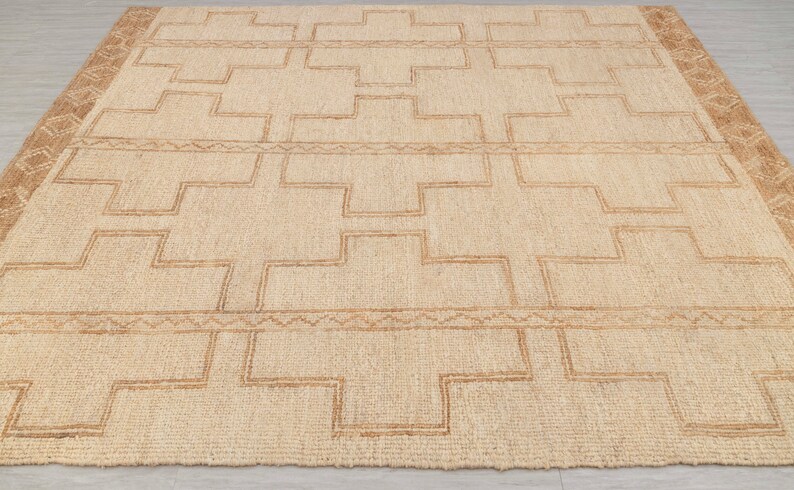 May include: A natural fiber rug with a geometric pattern in a light brown color. The rug has a woven texture and a subtle sheen.