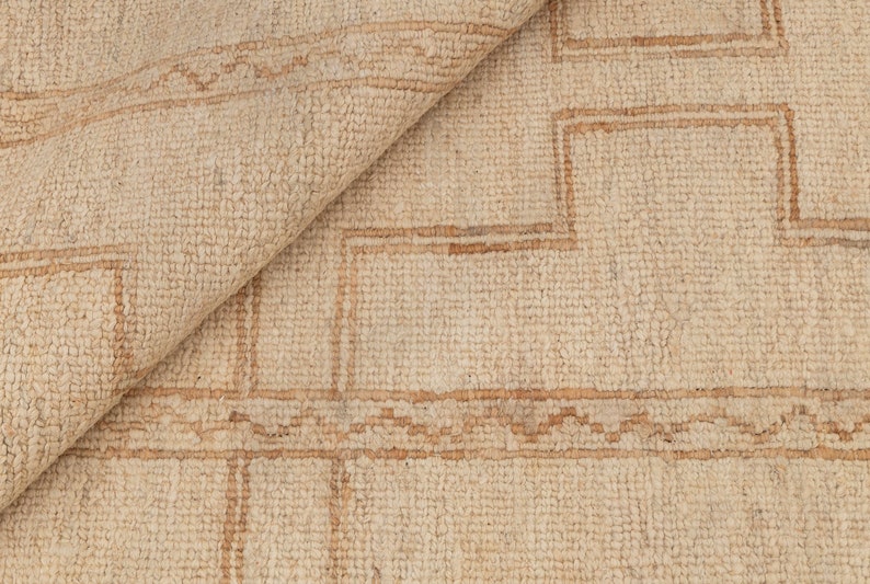 May include: A close-up of a cream-colored rug with a geometric pattern in a darker brown shade. The rug has a woven texture and a subtle sheen.