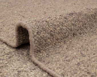 May include: Close-up of a rolled-up, textured rug in shades of beige and brown. The rug's surface has a nubby, speckled appearance, with a visible edge and a rolled section revealing the underside. The rug appears to be made of a thick, durable material.
