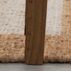 May include: Close-up of a natural fiber rug with a white stripe. A wooden ruler is placed next to the rug to show the texture and weave.