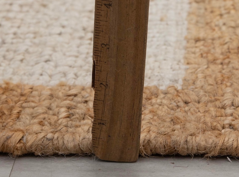 May include: Close-up of a natural fiber rug with a white stripe. A wooden ruler is placed next to the rug to show the texture and weave.