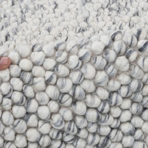 May include: Close-up of a white and gray textured rug made of small, round balls of wool. The rug has a soft, fluffy texture.