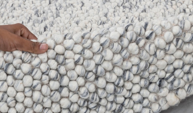 May include: Close-up of a white and gray textured rug made of small, round balls of wool. The rug has a soft, fluffy texture.