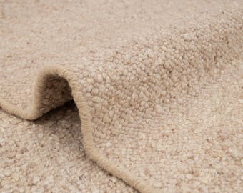 May include: Close-up of a beige rug with a textured, looped pile. The rug's edge is visible, showing a neatly finished border. The neutral color and thick texture suggest a cozy, durable floor covering.