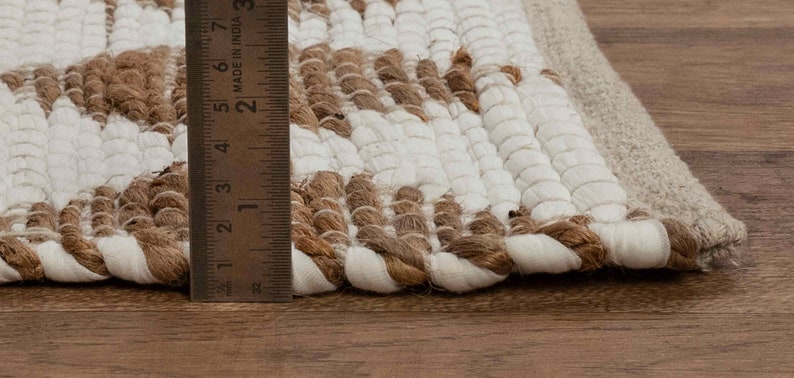 May include: Close-up of a white and brown woven rug with a fringe edge. A ruler is used to show the rug is approximately 2 inches thick.
