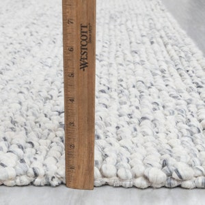 May include: Close-up of a white and gray textured rug with a wooden ruler measuring 7 inches. The rug has a fluffy, looped texture.