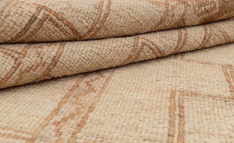 May include: A close-up of a beige and brown woven rug with a geometric pattern. The rug is folded in half, showing the texture and detail of the weave.