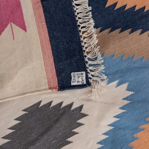 May include: A close-up of a woven rug with a geometric pattern in shades of blue, brown, and pink. The rug has a fringe border and a label that reads "8-238 DK 4111".