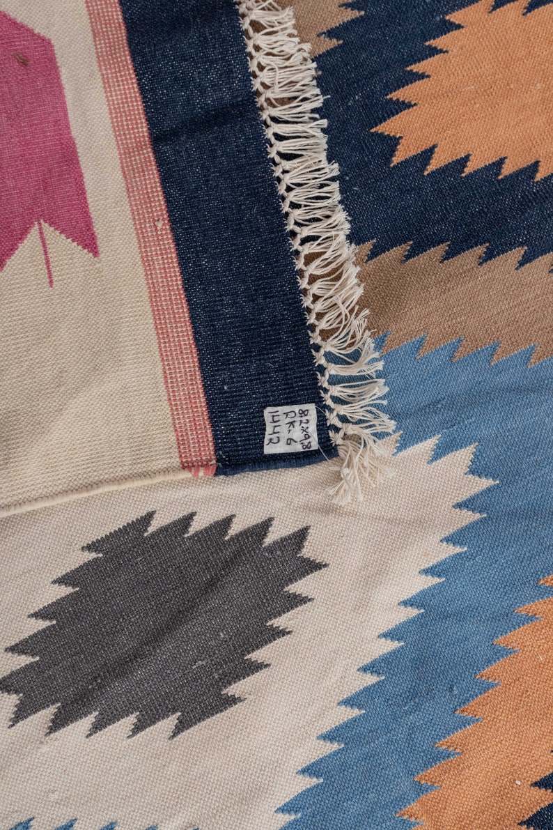May include: A close-up of a woven rug with a geometric pattern in shades of blue, brown, and pink. The rug has a fringe border and a label that reads "8-238 DK 4111".