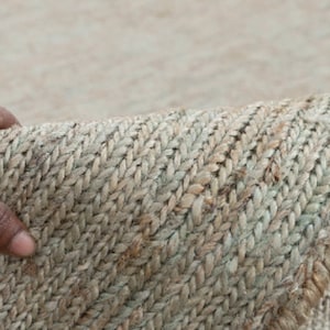 May include: Close-up of a natural fiber rug with a woven texture. The rug is a light beige color with a subtle green hue. The rug is folded in half and a hand is holding the edge of the rug.