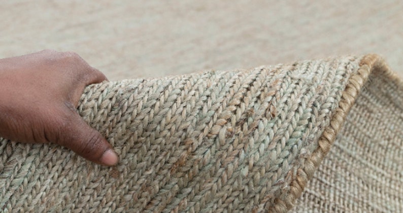 May include: Close-up of a natural fiber rug with a woven texture. The rug is a light beige color with a subtle green hue. The rug is folded in half and a hand is holding the edge of the rug.