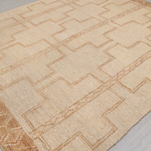 May include: A beige rug with a geometric pattern in a darker beige color. The rug has a border with a more intricate pattern.