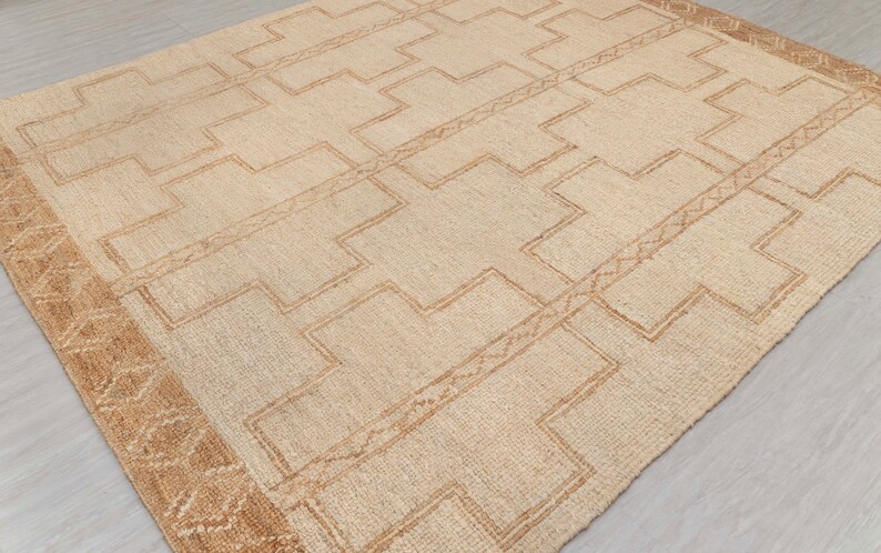 May include: A beige rug with a geometric pattern in a darker beige color. The rug has a border with a more intricate pattern.