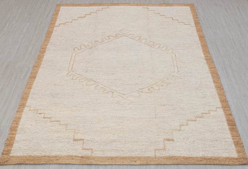 May include: A beige rug with a geometric pattern in a lighter beige color. The rug has a brown border.