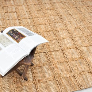 Puede incluir: Una alfombra grande y rectangular de yute con un patrón tejido en forma de cuadrícula. Un libro abierto descansa sobre un pequeño taburete de madera, mostrando imágenes coloridas. El color y la textura naturales de la alfombra crean un ambiente cálido.
