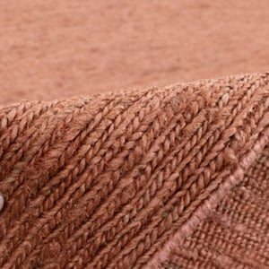 May include: Close-up of a hand holding a woven rug with a textured, reddish-brown surface. The rug has a subtle pattern of diagonal lines.