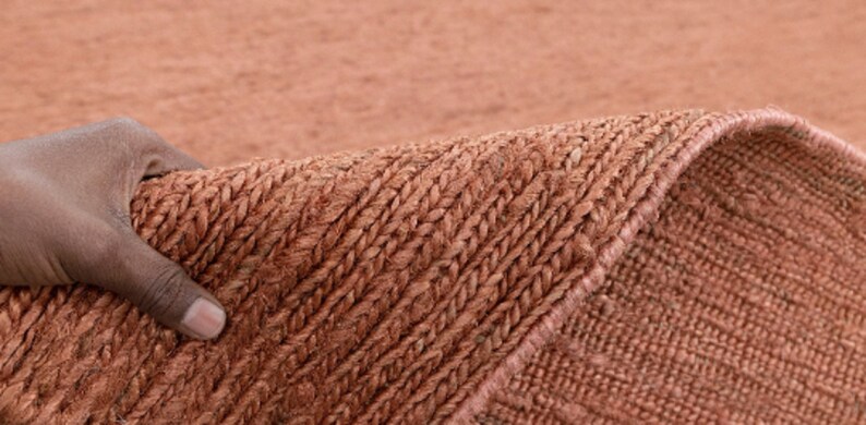 May include: Close-up of a hand holding a woven rug with a textured, reddish-brown surface. The rug has a subtle pattern of diagonal lines.