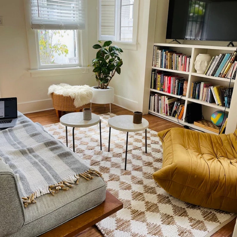 May include: A living room with a white and brown patterned rug, two round side tables, a white couch with a gray throw blanket, a white wicker ottoman with a white faux fur throw, a potted plant, a bookshelf filled with books, a yellow beanbag chair, and a television.