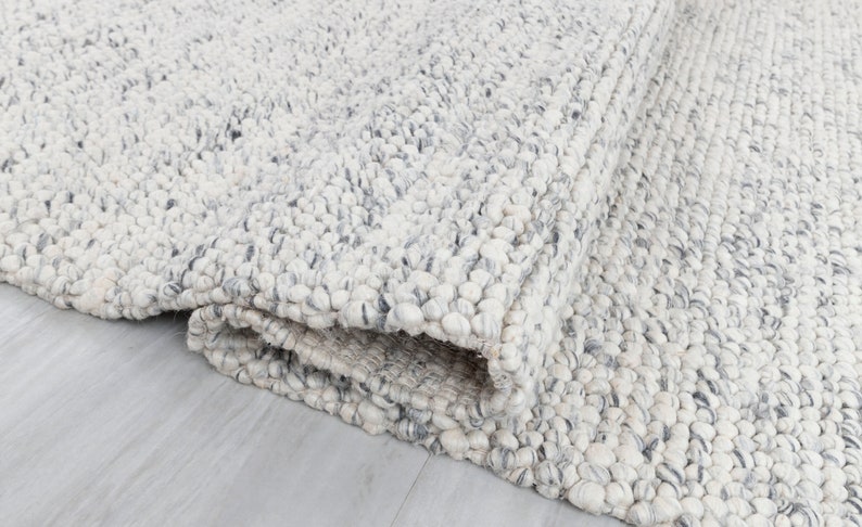 May include: A close-up of a white and gray textured rug with a fluffy, nubby texture. The rug is folded in half, showing the underside of the rug.