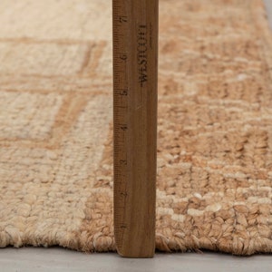 May include: A close-up of a natural fiber rug with a woven pattern. The rug is beige and brown in color and has a frayed edge. A wooden ruler is placed next to the rug for scale.