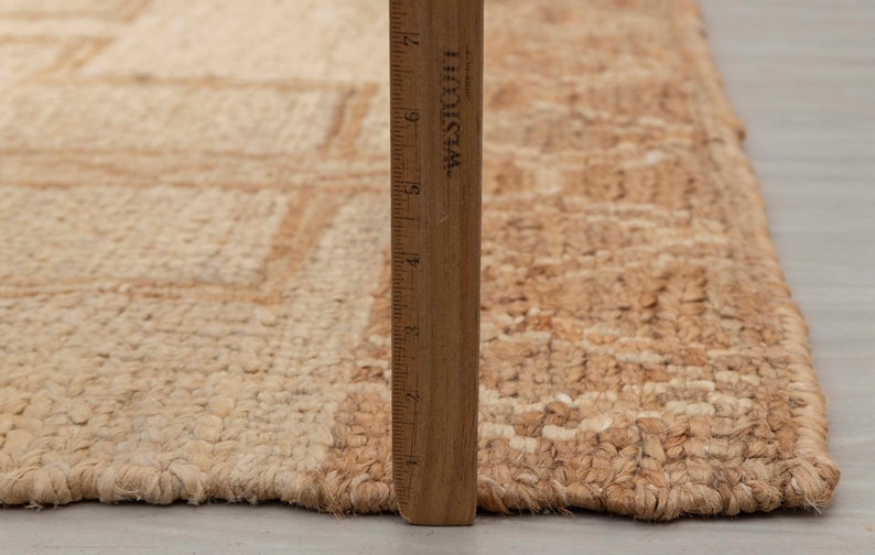 May include: A close-up of a natural fiber rug with a woven pattern. The rug is beige and brown in color and has a frayed edge. A wooden ruler is placed next to the rug for scale.
