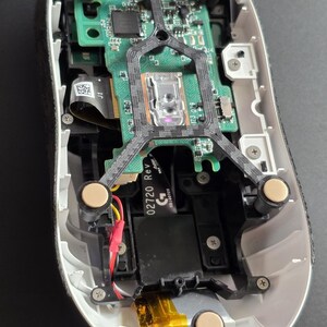 May include: The internal components of a computer mouse with a white outer shell, a green circuit board, a black plastic frame, and four circular beige feet. A black ribbon cable is visible with the text 02720 Rev and a G logo printed on it.