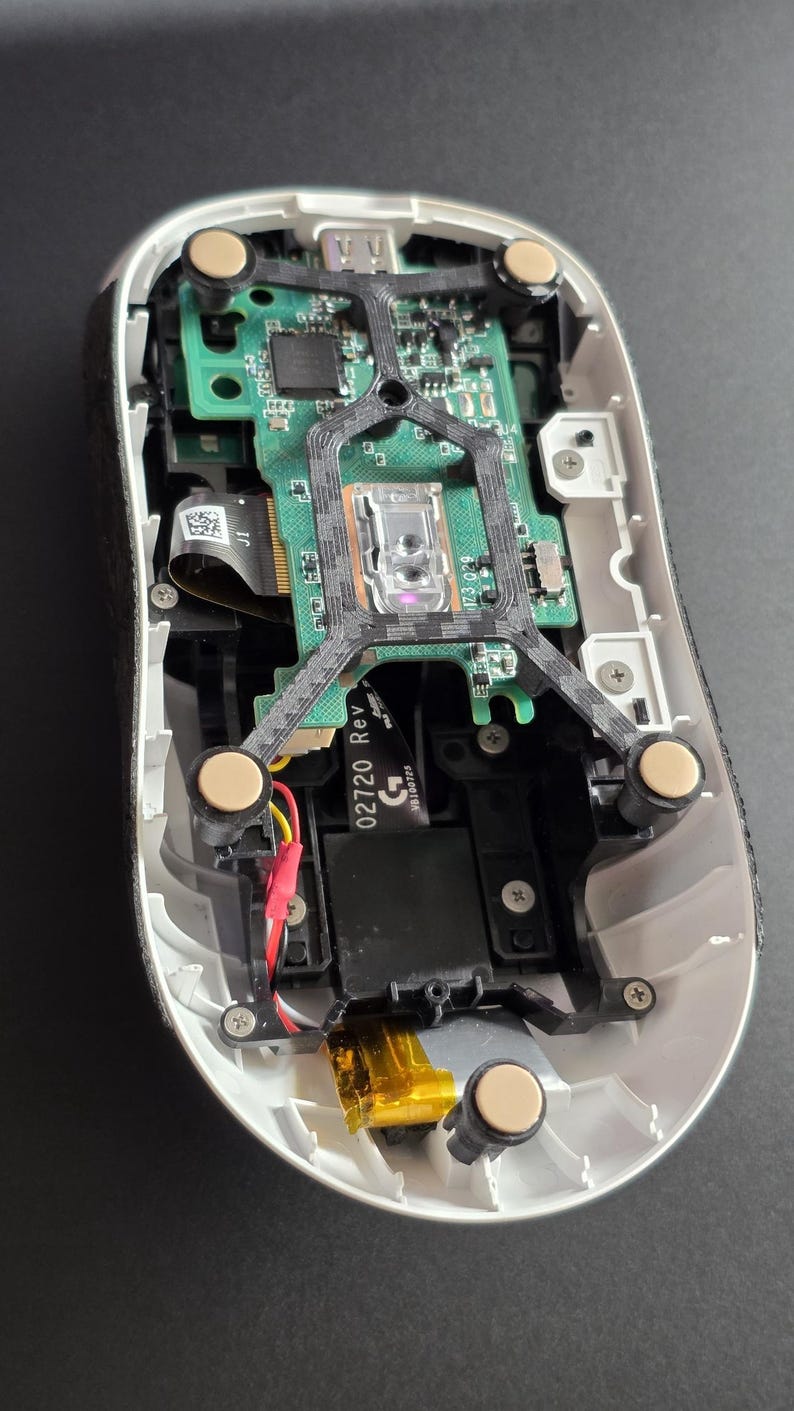 May include: The internal components of a computer mouse with a white outer shell, a green circuit board, a black plastic frame, and four circular beige feet. A black ribbon cable is visible with the text 02720 Rev and a G logo printed on it.