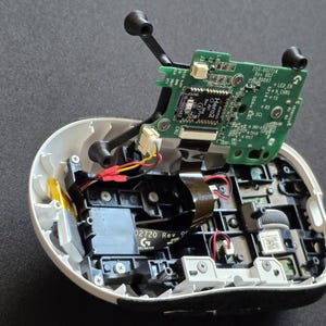 May include: An opened computer mouse with its white outer shell partially removed, revealing internal black plastic components, a grey scroll wheel, and a green circuit board labeled Hero2, connected by thin red, yellow, and black wires.
