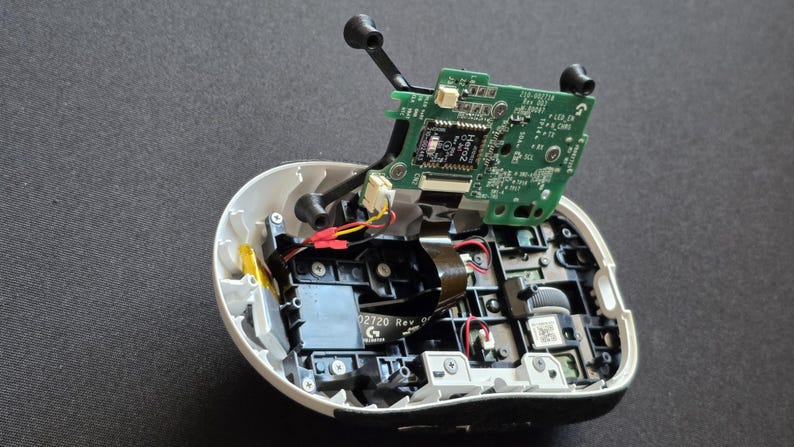 May include: An opened computer mouse with its white outer shell partially removed, revealing internal black plastic components, a grey scroll wheel, and a green circuit board labeled Hero2, connected by thin red, yellow, and black wires.