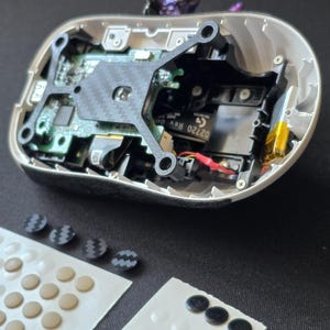 May include: A disassembled computer mouse with an exposed green circuit board, a black carbon fiber-patterned internal frame, and internal wiring. Included are two adhesive sheets containing multiple small, circular, beige and black replacement feet.