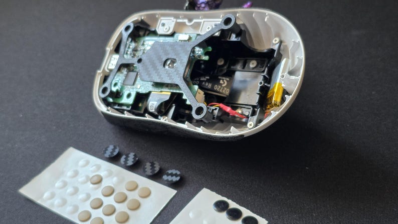 May include: A disassembled computer mouse with an exposed green circuit board, a black carbon fiber-patterned internal frame, and internal wiring. Included are two adhesive sheets containing multiple small, circular, beige and black replacement feet.