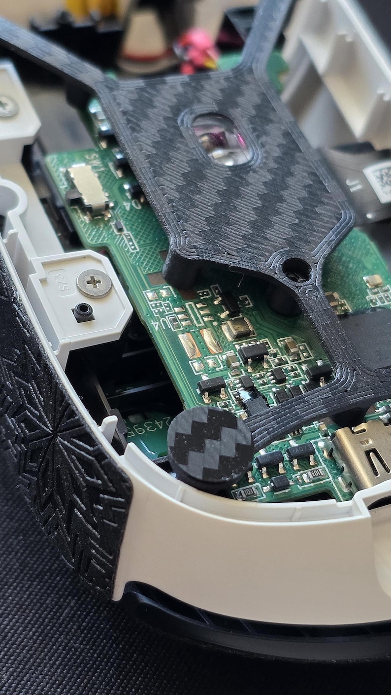 May include: An internal view of a disassembled electronic device featuring a green circuit board with various microchips and components. A black 3D-printed frame with a carbon fiber-like texture covers parts of the board, alongside white plastic housing.