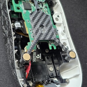 May include: The internal components of a computer mouse with a white outer shell and black textured side panels. A green circuit board is visible, featuring a central carbon fiber pattern support structure, gold-colored contact points, and a yellow battery.