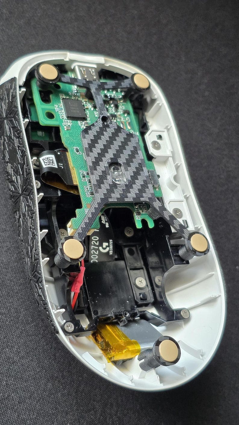 May include: The internal components of a computer mouse with a white outer shell and black textured side panels. A green circuit board is visible, featuring a central carbon fiber pattern support structure, gold-colored contact points, and a yellow battery.