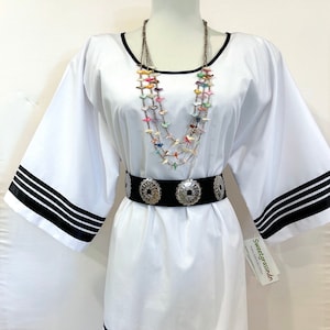 May include: White tunic with black trim around the neckline, sleeves, and hem. The tunic is belted with a black belt with silver conchos. A multi-colored bird necklace is worn over the tunic. The tag reads "Sweetgrassdn Native American Crafts".