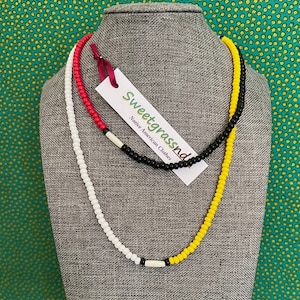 Può includere: Due collane di perline esposte su un manichino grigio. Una collana è bianca, rossa e nera, con un'etichetta che recita "Sweetgrassnd Native American Clothes". L'altra collana è gialla e nera. Lo sfondo è un tessuto verde con motivi floreali gialli.