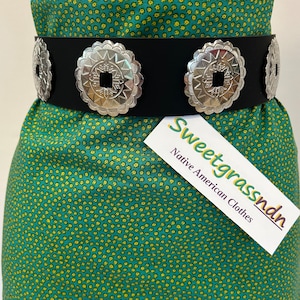 Puede incluir: Un cinturón de cuero negro con detalles de concho plateado. El cinturón se lleva sobre un vestido verde con estampado floral blanco. El texto "Sweetgrassndn Native American Clothes" es visible en una etiqueta blanca.