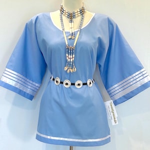 May include: A light blue tunic with wide sleeves and white trim. The tunic is paired with a beaded necklace and a belt with silver and black accents. The sleeves have a white stripe design. The tunic is displayed on a mannequin.