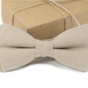 Può includere: Un papillon in lino beige è esposto davanti a una confezione regalo. Il papillon, al centro, mette in risalto il suo tessuto strutturato e la sua forma classica. La confezione regalo è avvolta in carta marrone e legata con spago.